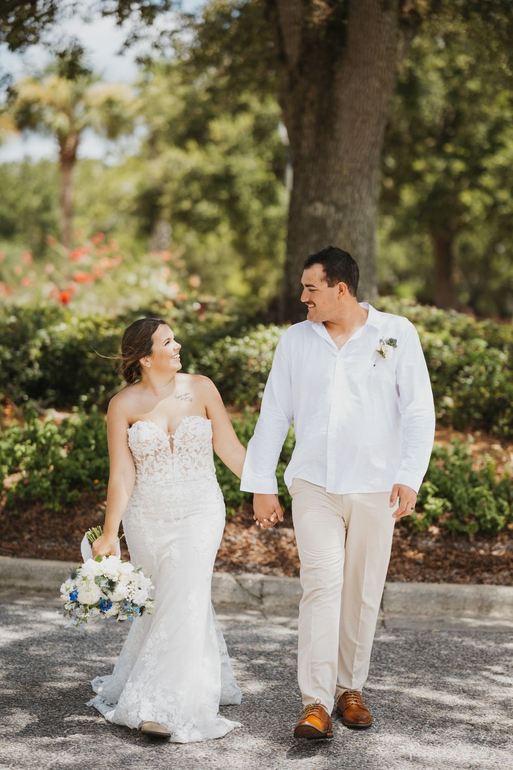 Bride and groom walk hand-in-hand outdoors, she in lace gown with bouquet, he in white shirt and khakis.