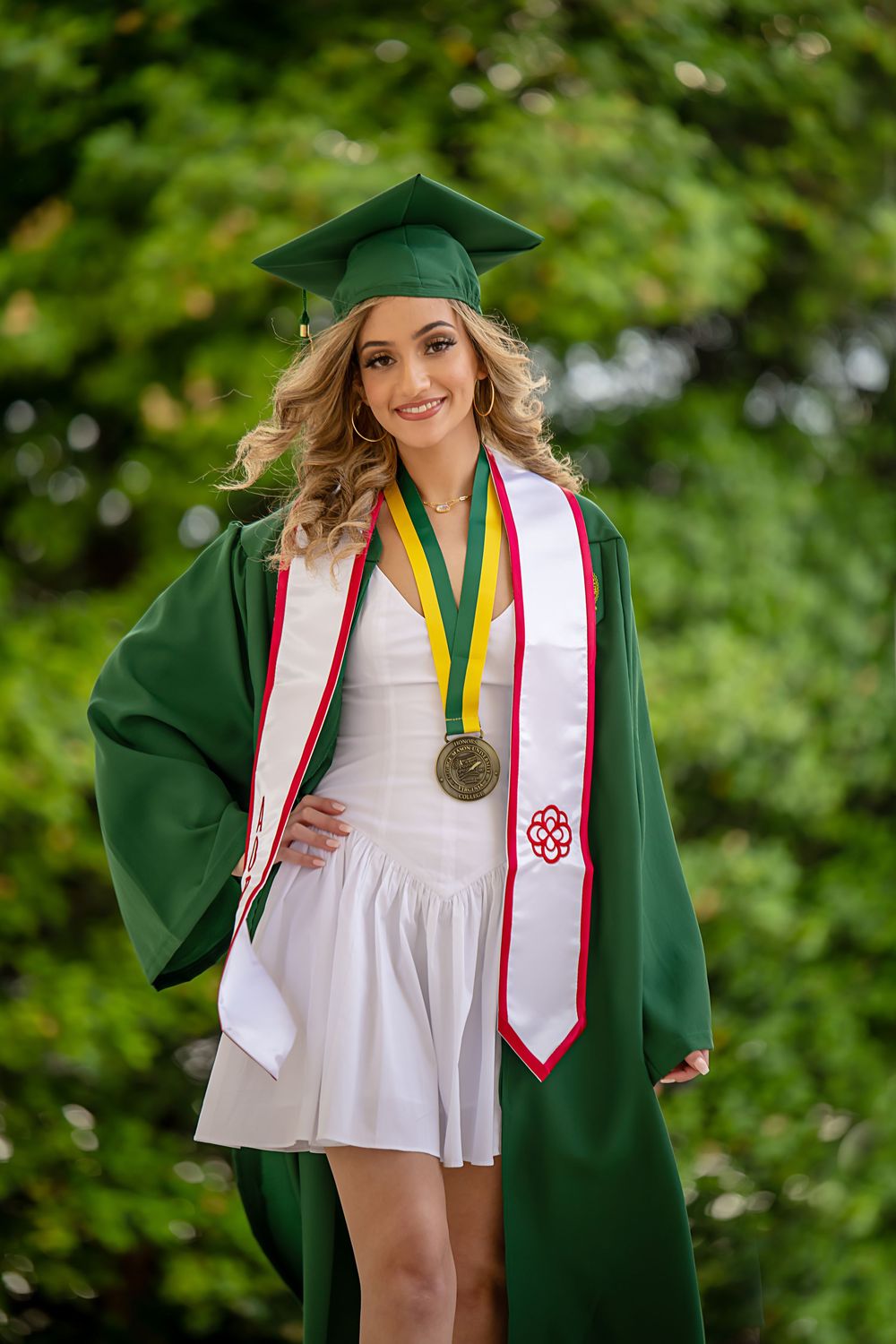 Graduate in green cap and gown with colorful stoles against lush green background.
