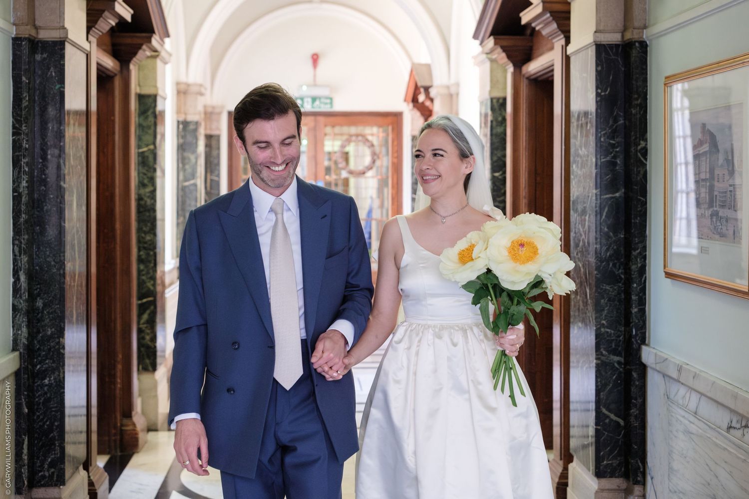Happy couple walking through elegant marble hallway after wedding ceremony with yellow floral bouquet.