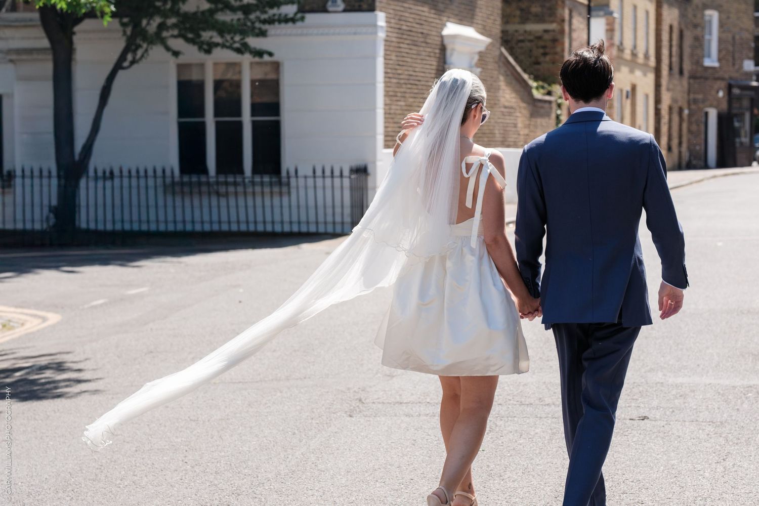 Newlywed couple holding hands while walking down a sunny street, with bride in short dress and flowing veil.