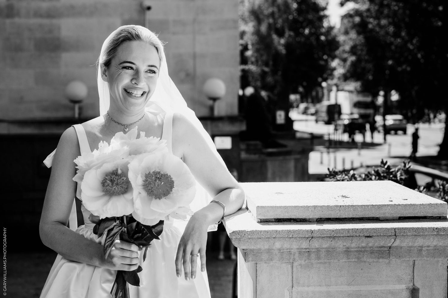 A bride in a white dress holds a large sunflower bouquet while standing beside a stone ledge outdoors.