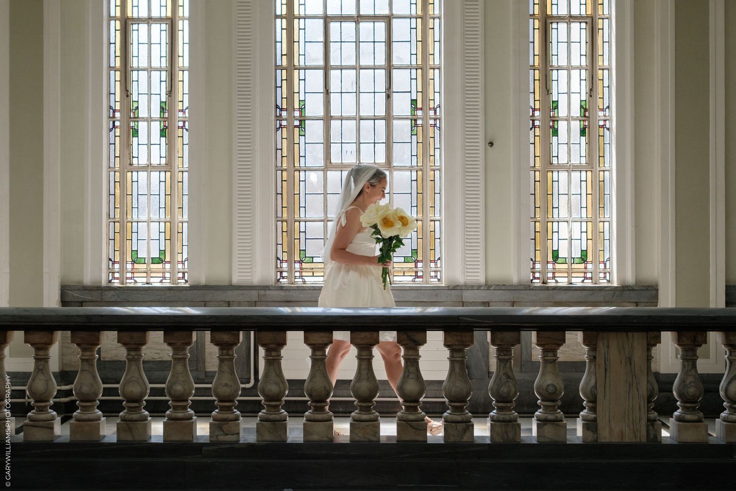 A person in a white dress holds a sunflower bouquet while standing by ornate windows in an elegant interior hallway.