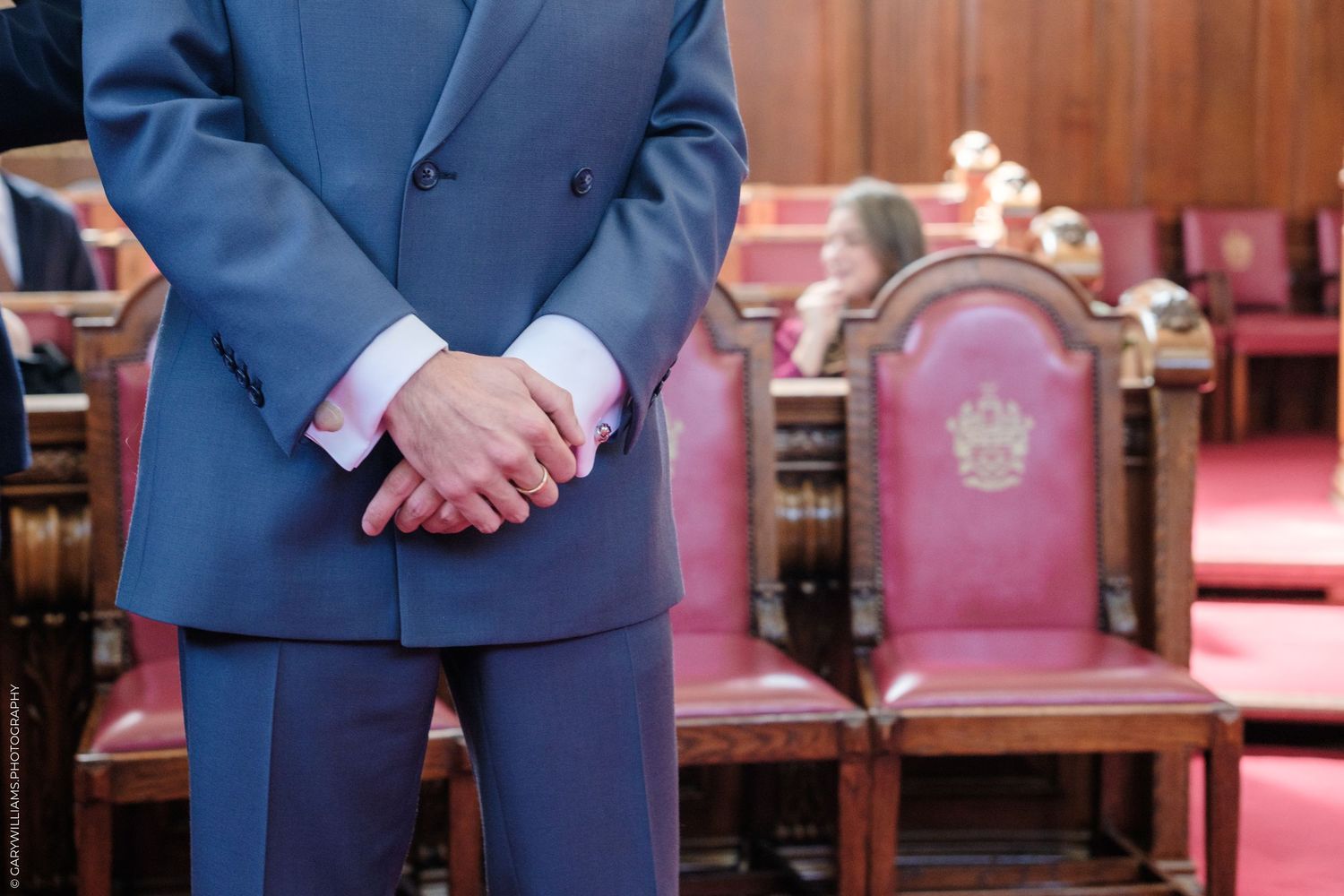 Person in navy blue suit standing next to ornate red chairs with decorative wooden carvings in formal setting.