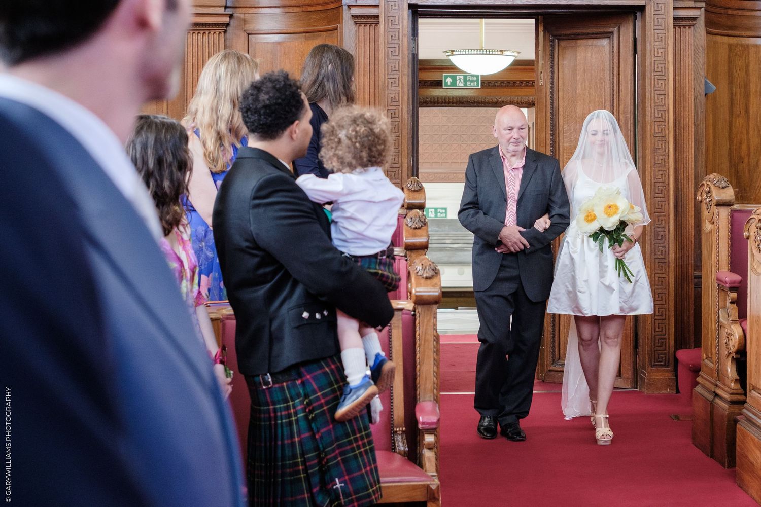A church wedding ceremony shows a small gathering of guests in formal attire including kilts watching from the aisle.