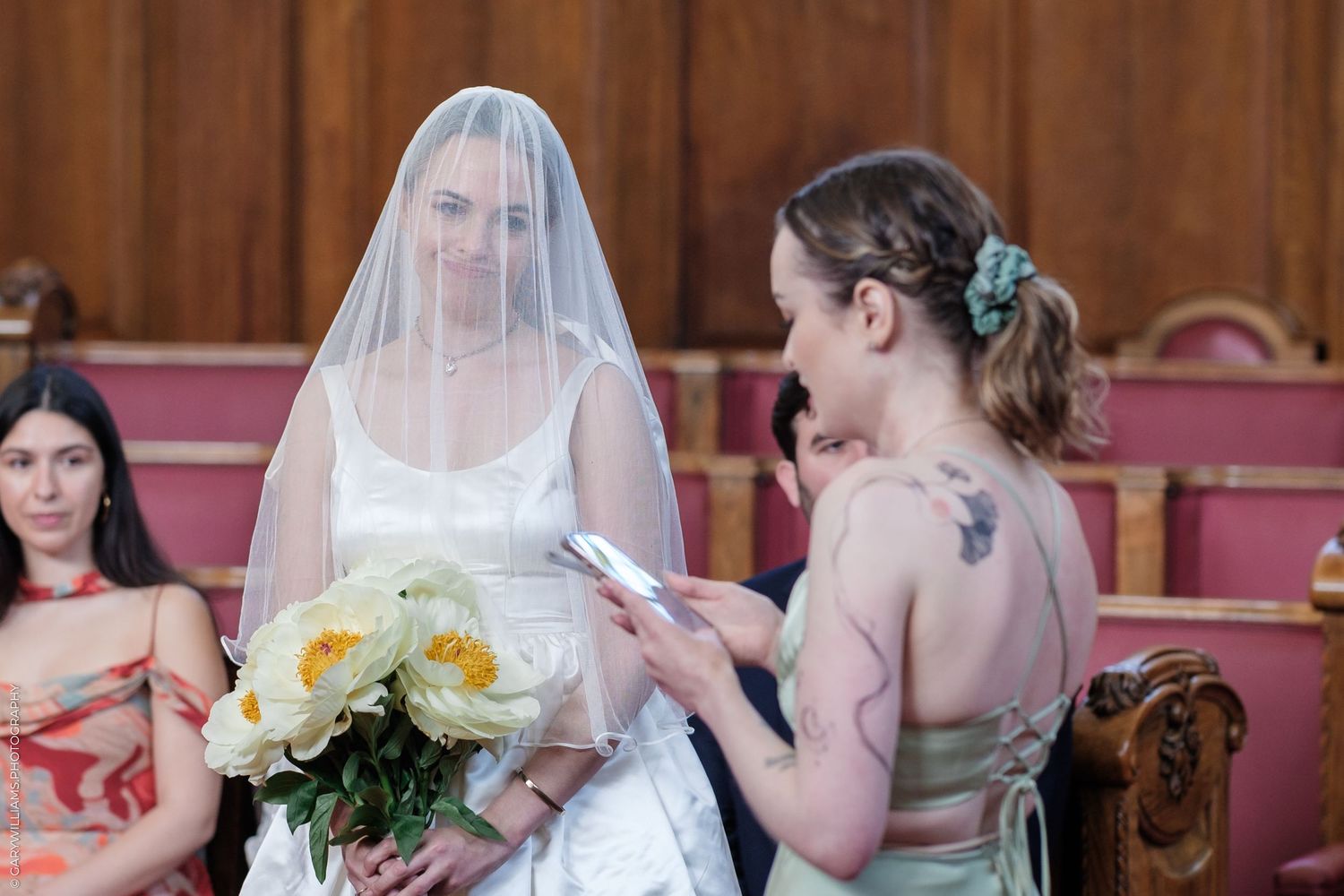 Bride in white dress with veil holding yellow bouquet stands with bridesmaid in church pews during wedding ceremony.
