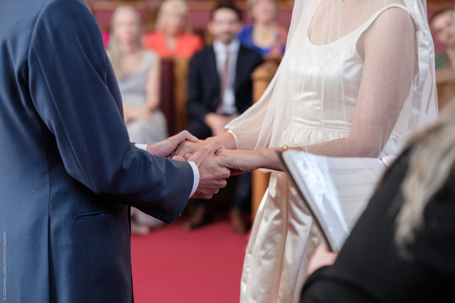 Close-up of hands exchanging wedding rings during a ceremony on a red carpet with guests in background.
