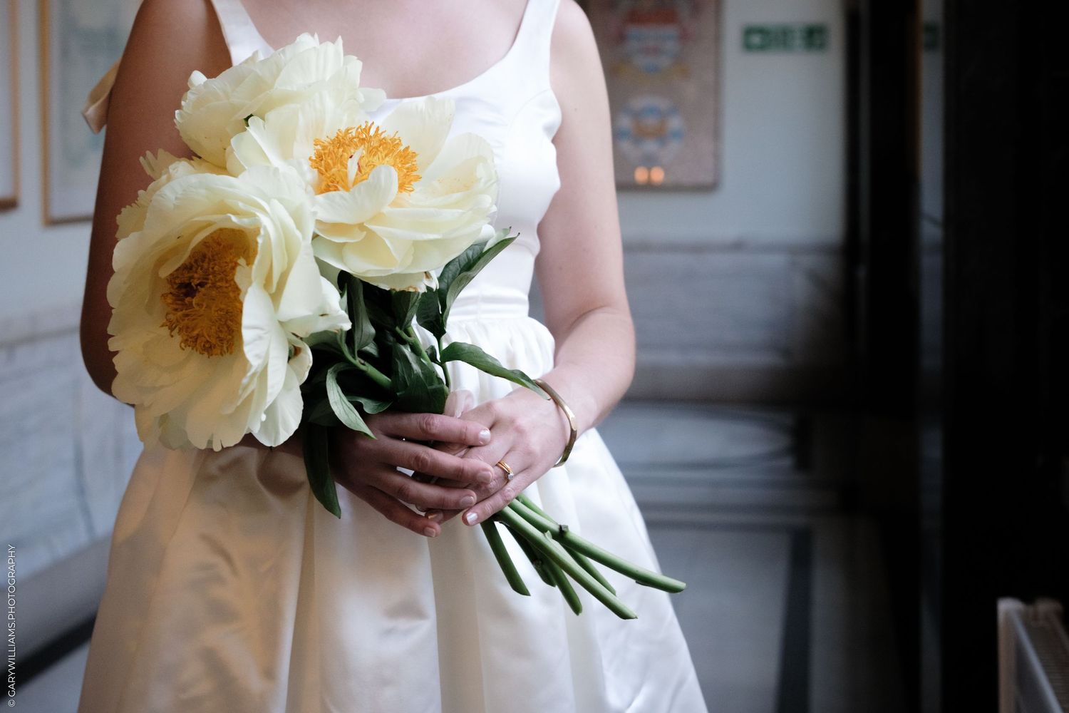 A bouquet of large cream-colored peony flowers with orange centers held against a white dress in soft lighting.