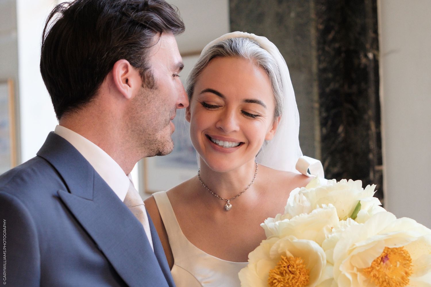Newlywed couple shares an intimate moment with white floral bouquet against dark marble background.