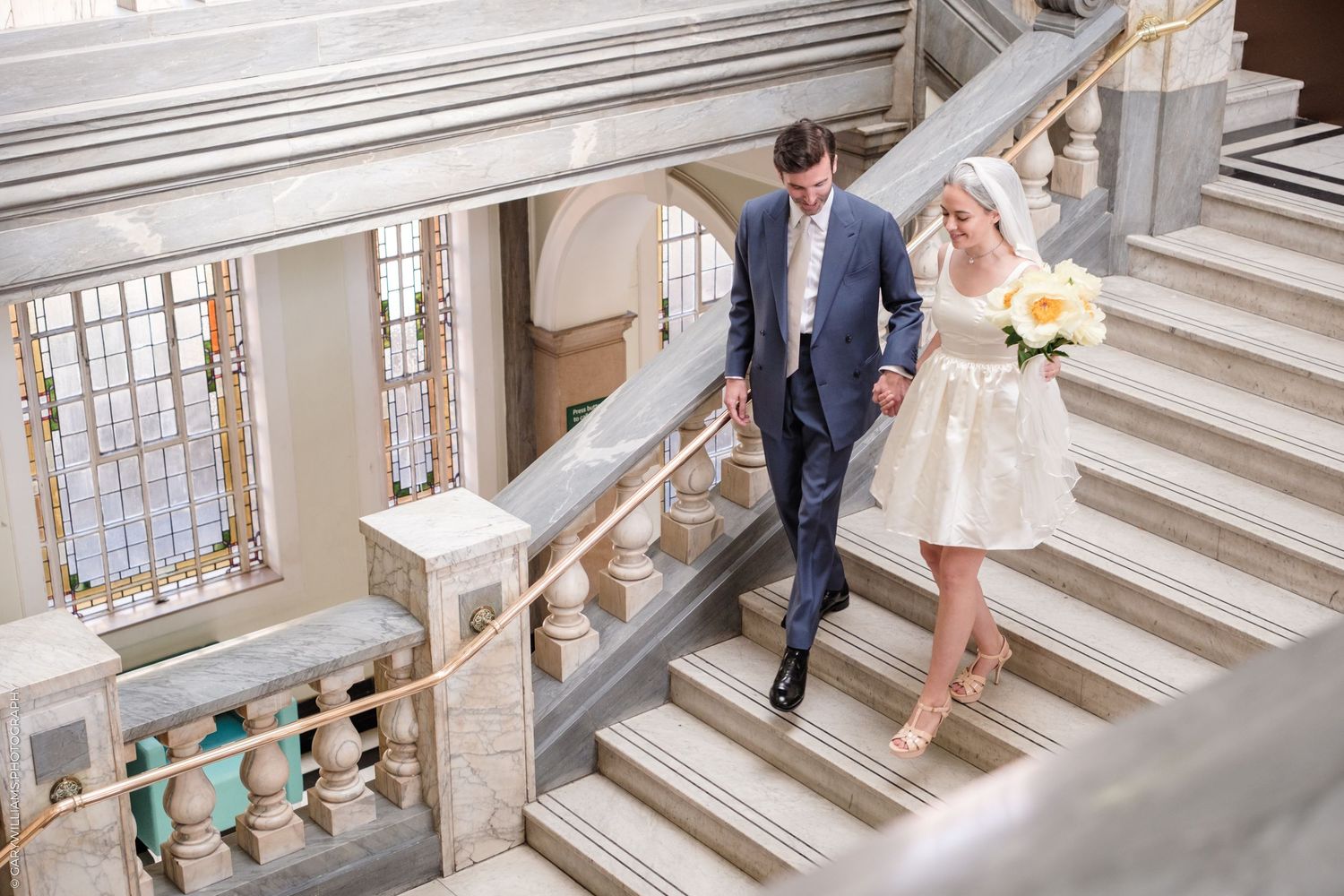 A couple in formal attire walking down an elegant architectural staircase in a grand interior setting.