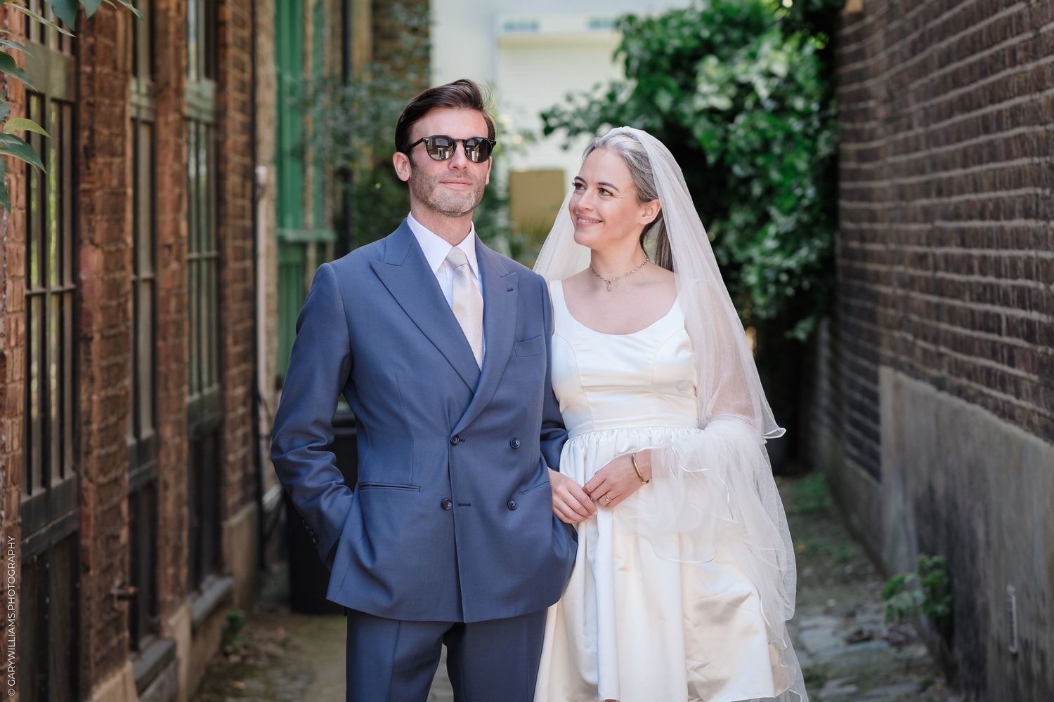 A bride in a white lace dress and veil stands with a groom wearing a gray suit in a brick alleyway.