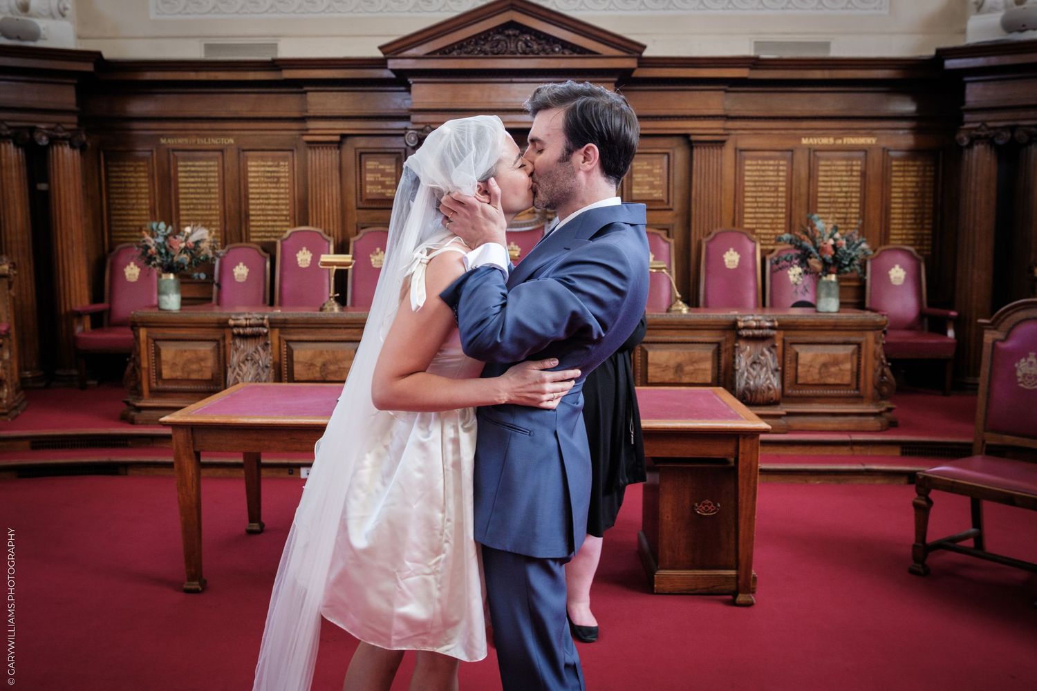 A couple shares a tender kiss in a traditional church setting with red pews and wooden paneling in the background.