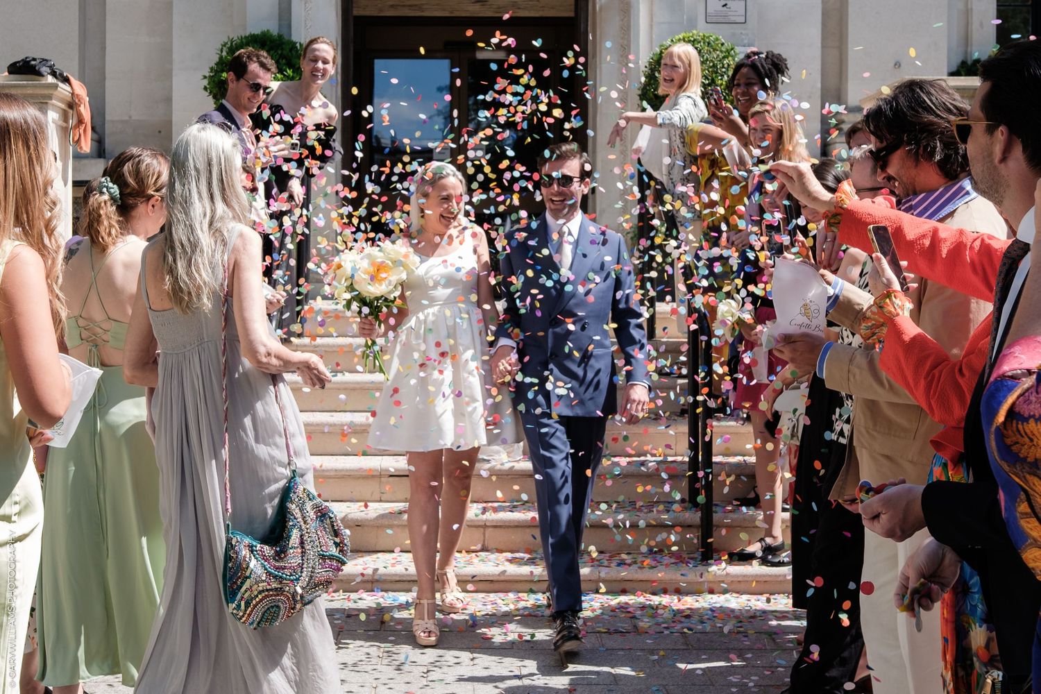 Wedding guests shower the happy newlyweds with white confetti as they exit down the stone church steps.