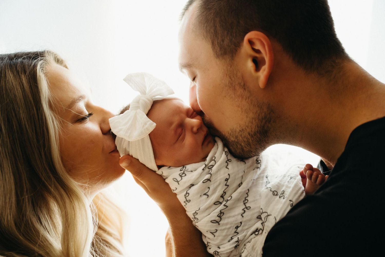 Parents sharing a tender moment with their newborn baby in warm sunlight through a window.