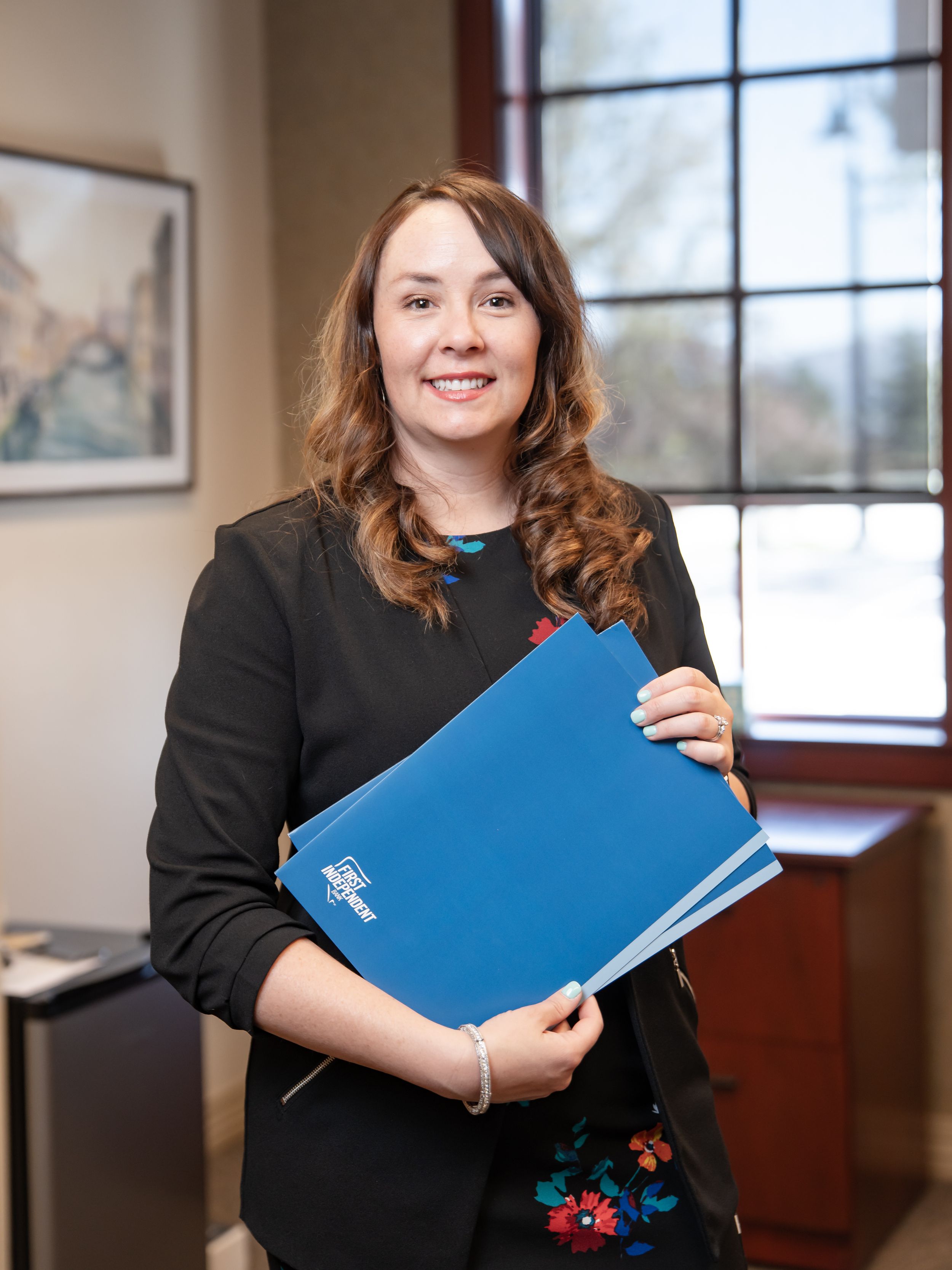 Professional in a black sweater with embroidered flowers holds a blue folder while standing in an office near a window.
