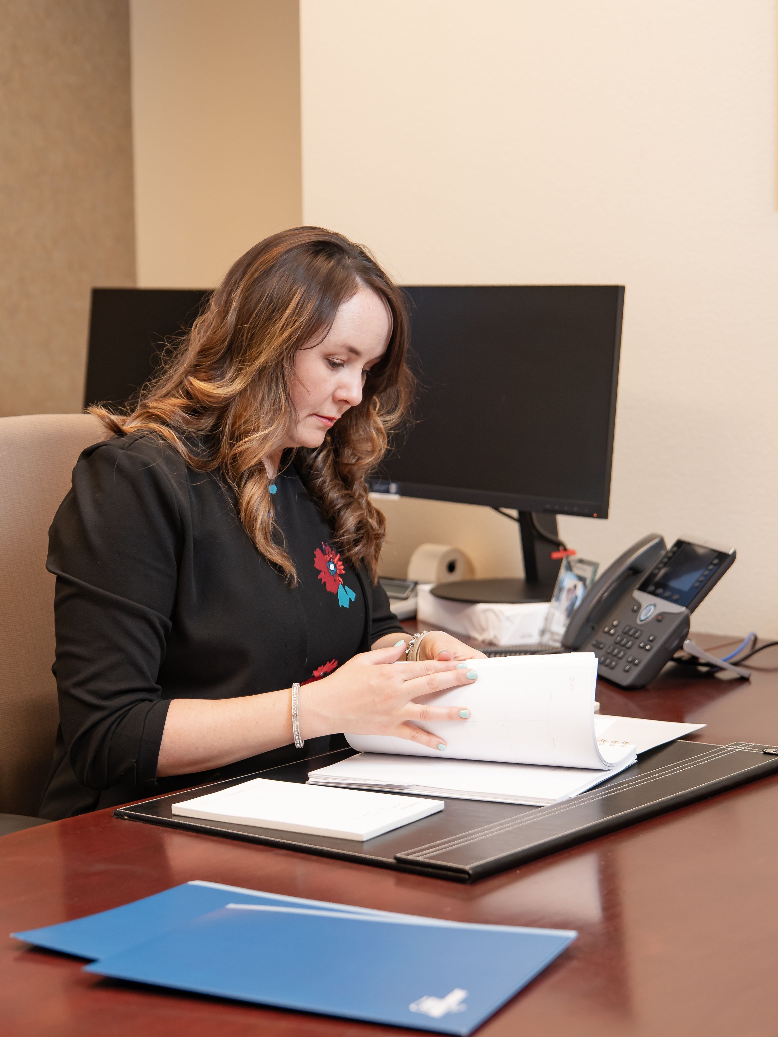 Professional working at office desk reviews documents and paperwork while seated at workstation with computer monitor.