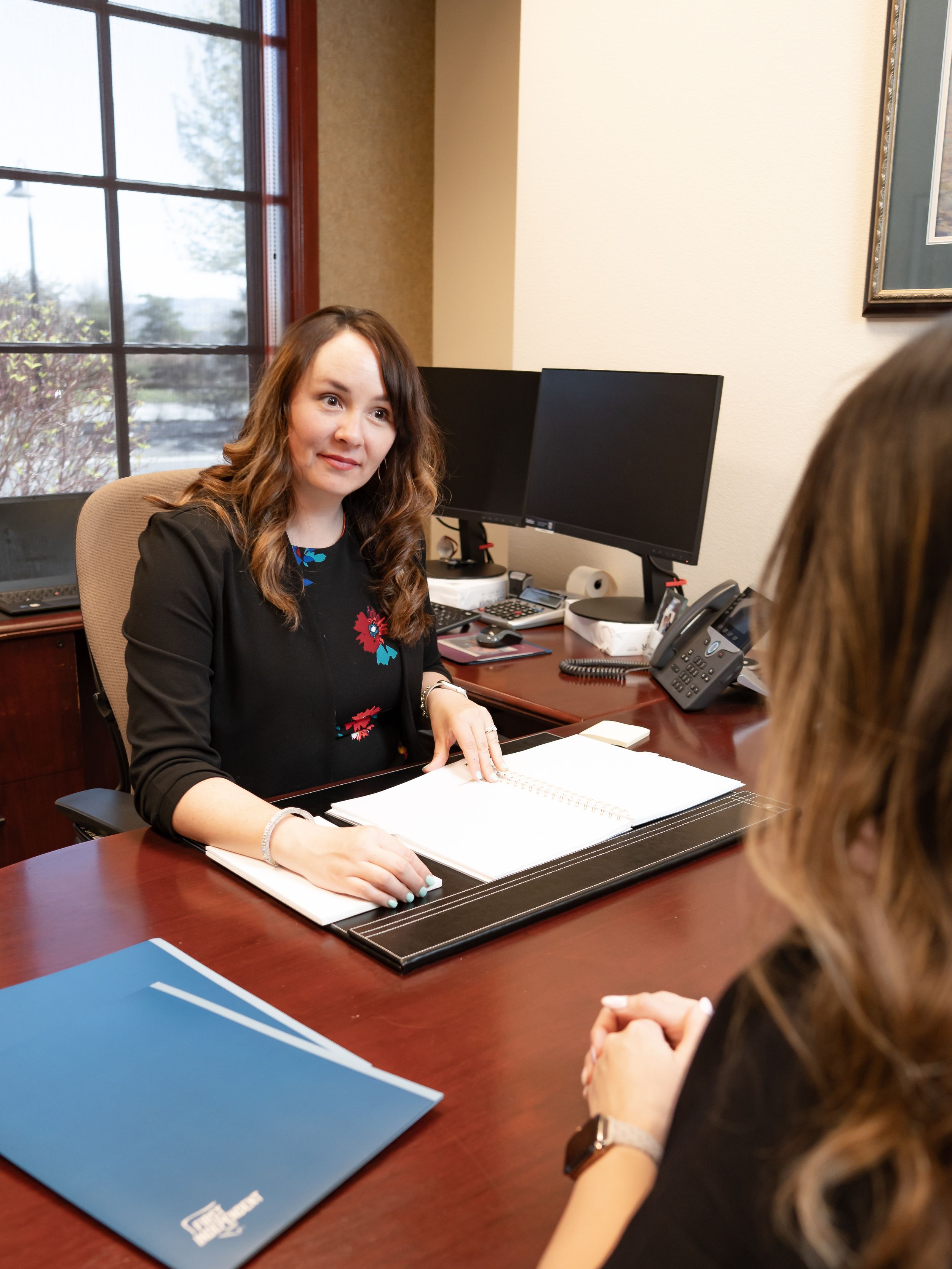 Professional meeting between two people at office desk with computer monitor and blue folder visible.