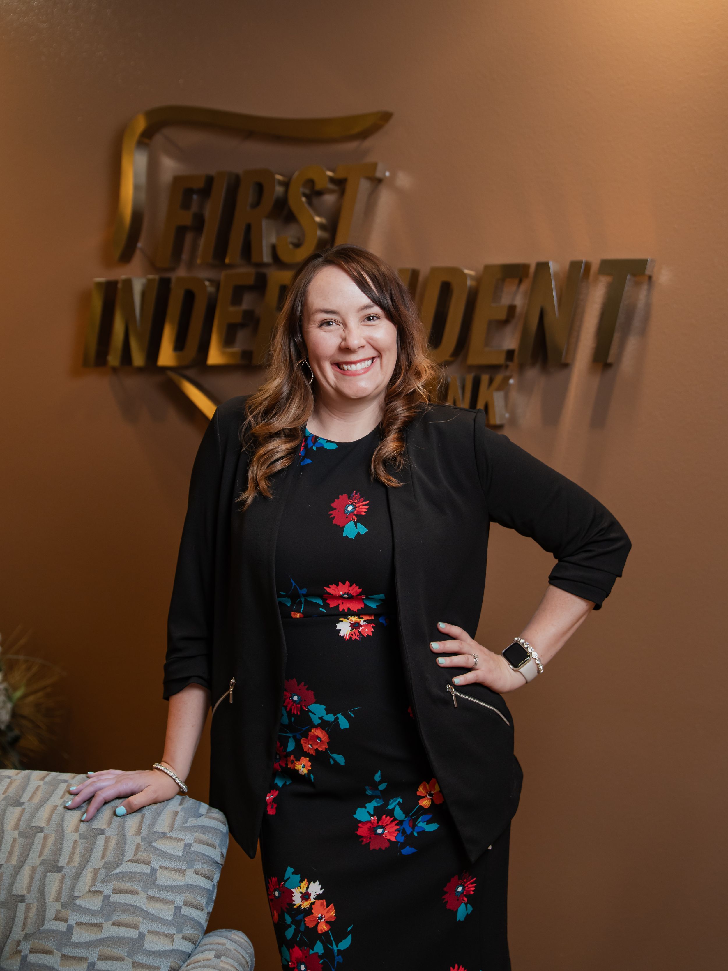 Professional portrait against brown office wall with First Independent logo showing person in black cardigan over floral dress.