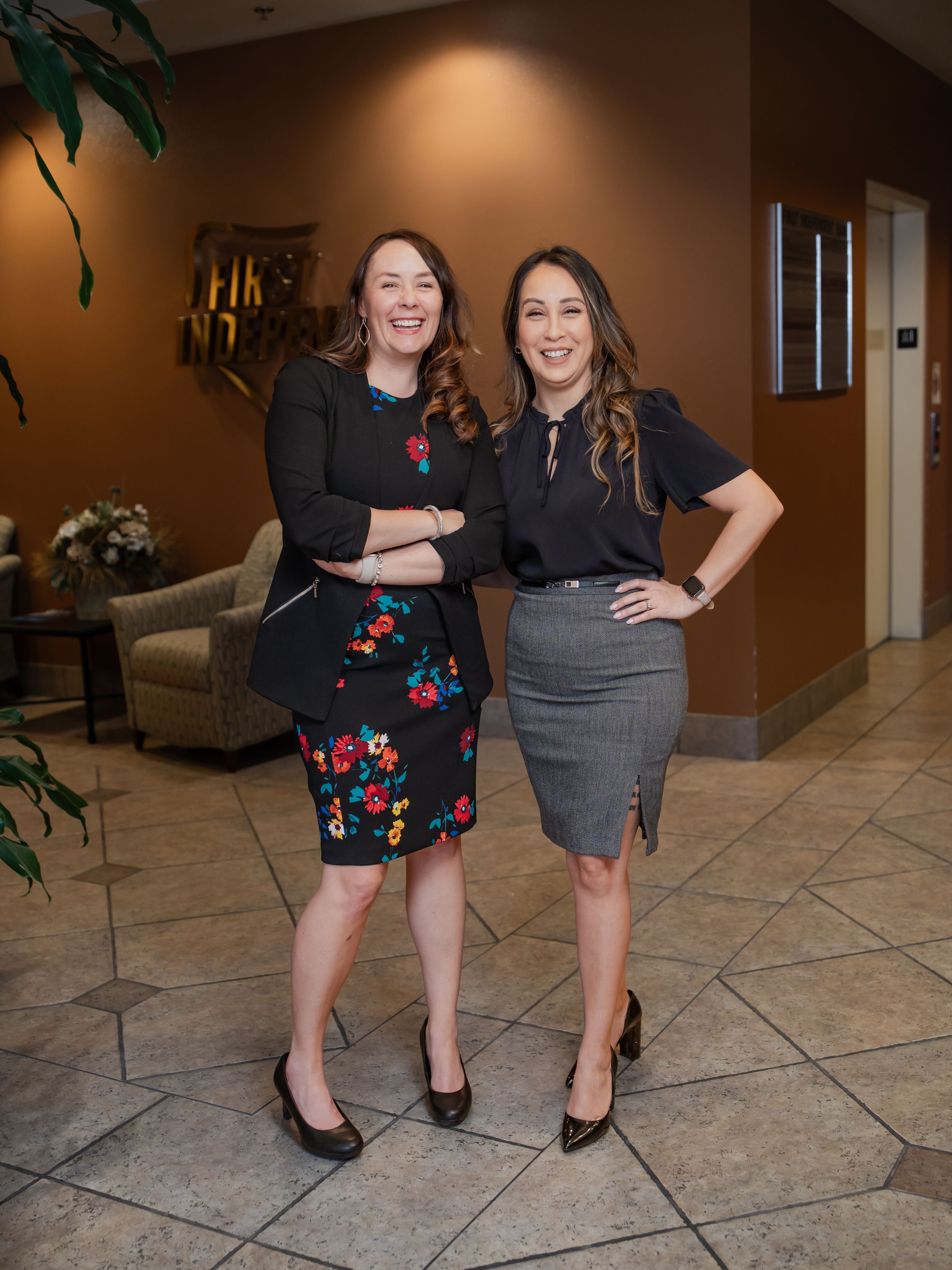 Two professionals in business attire pose together in an office lobby with tiled floors and brown walls.