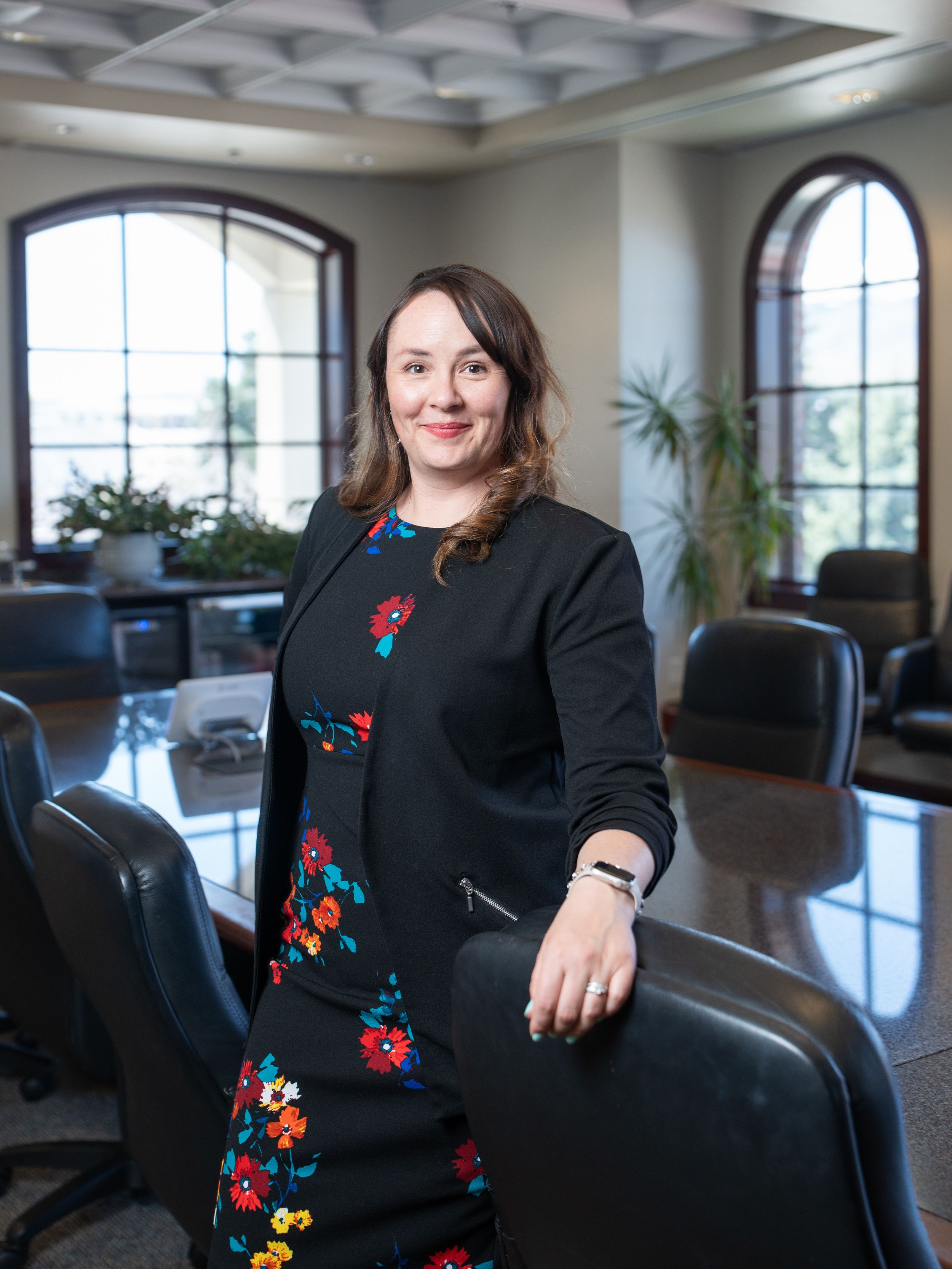Professional portrait in a modern office interior with arched windows, black furniture and decorative floral dress and cardigan.
