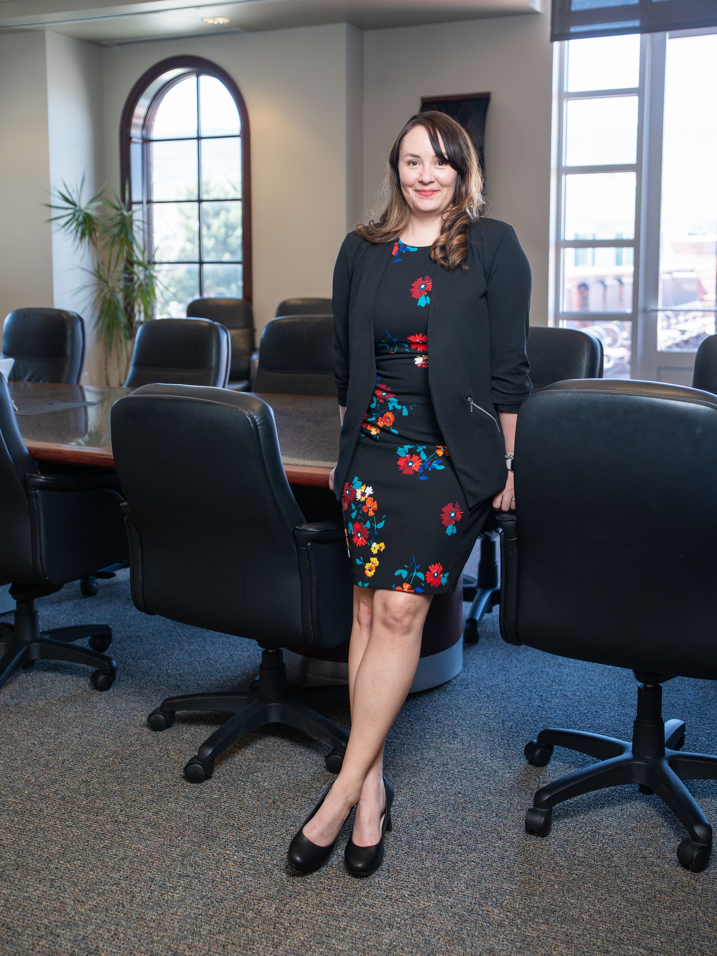 A businesswoman wearing a floral dress and black cardigan stands in a modern office conference room with arched windows.