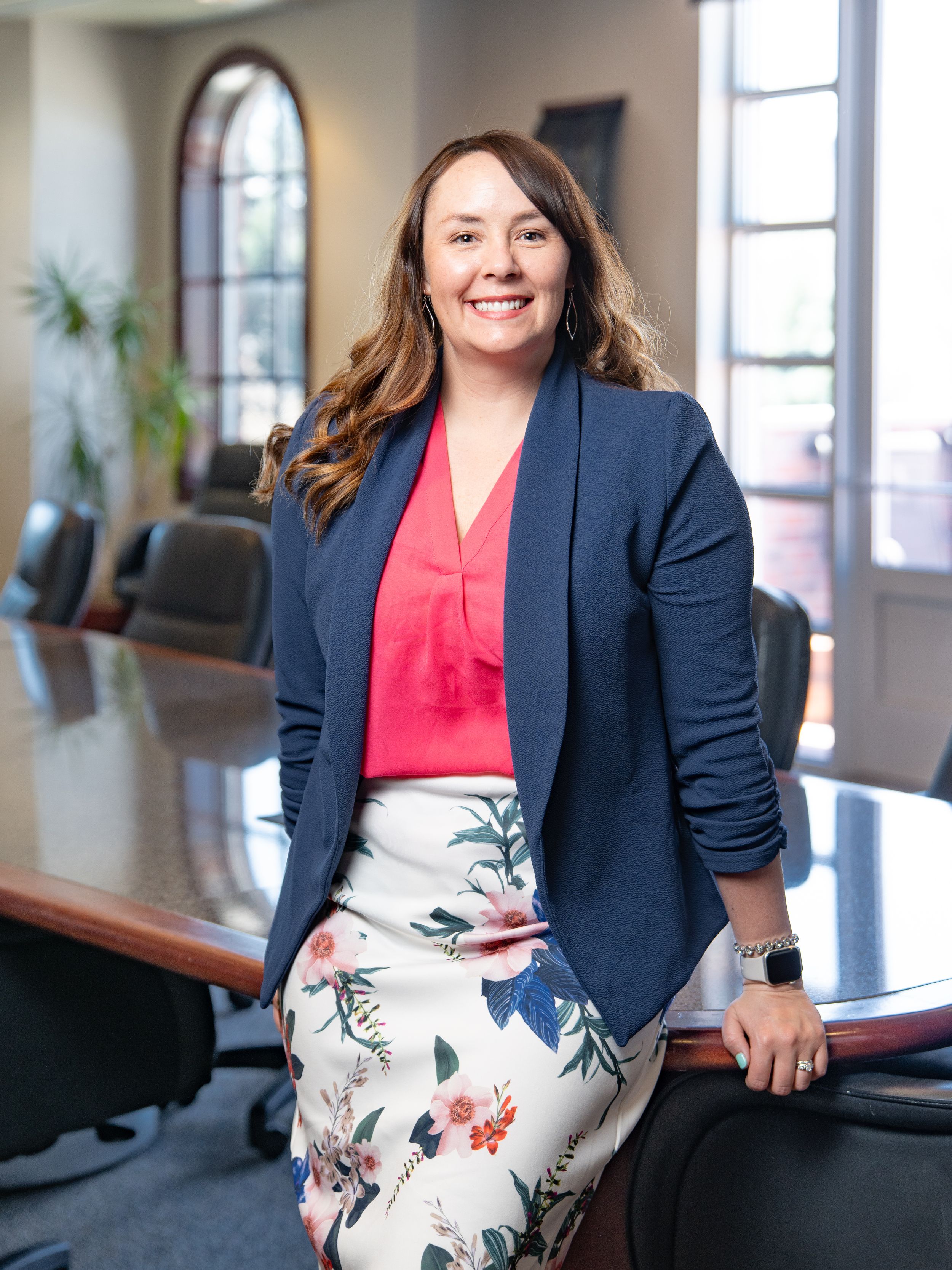 Professional wearing a navy blazer with coral top and floral pencil skirt stands confidently in a modern conference room.
