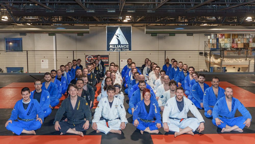 A large group of martial arts students in blue and white gis seated together on training mats at a gym facility.