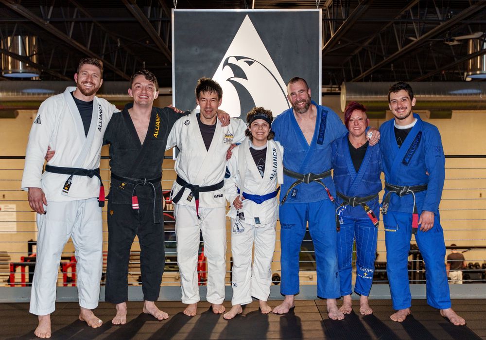 Group of martial artists in white and blue gis pose together before a black triangle flag on wooden wall.