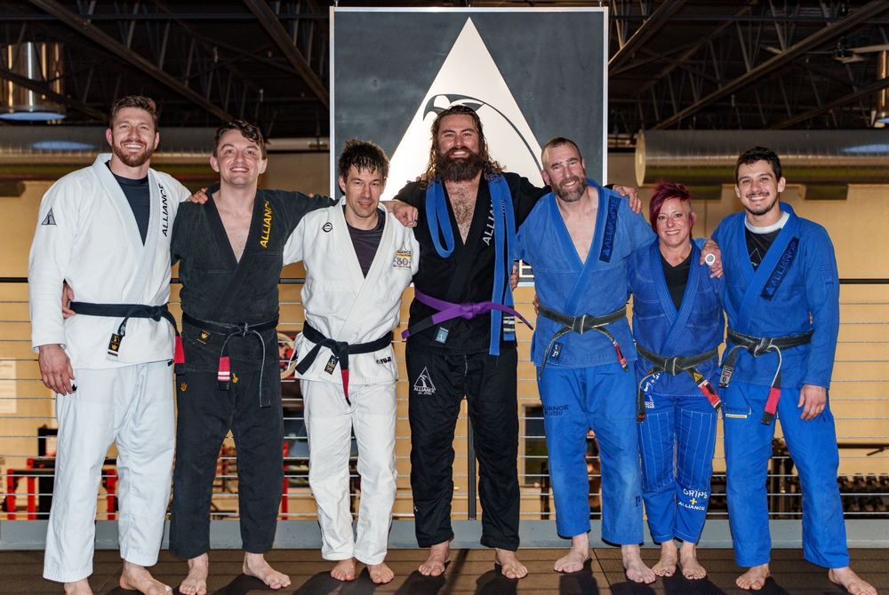 Martial arts practitioners wearing different colored gis pose together on training mats in a studio.