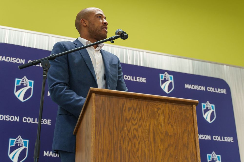 A speaker in a navy suit addresses an audience from a wooden podium with Madison College branding and logos visible.