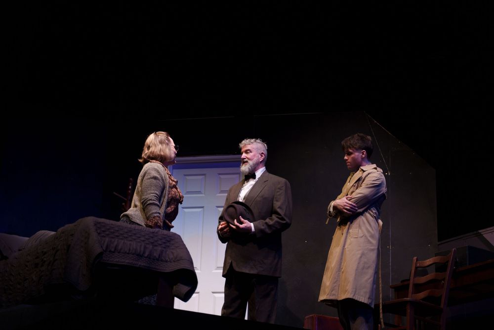 Three actors perform in period costumes on a dimly lit theater stage with vintage furniture and set pieces.
