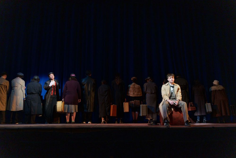 Theater performers stand on a dimly lit stage with vintage suitcases in period costumes during a dramatic scene.