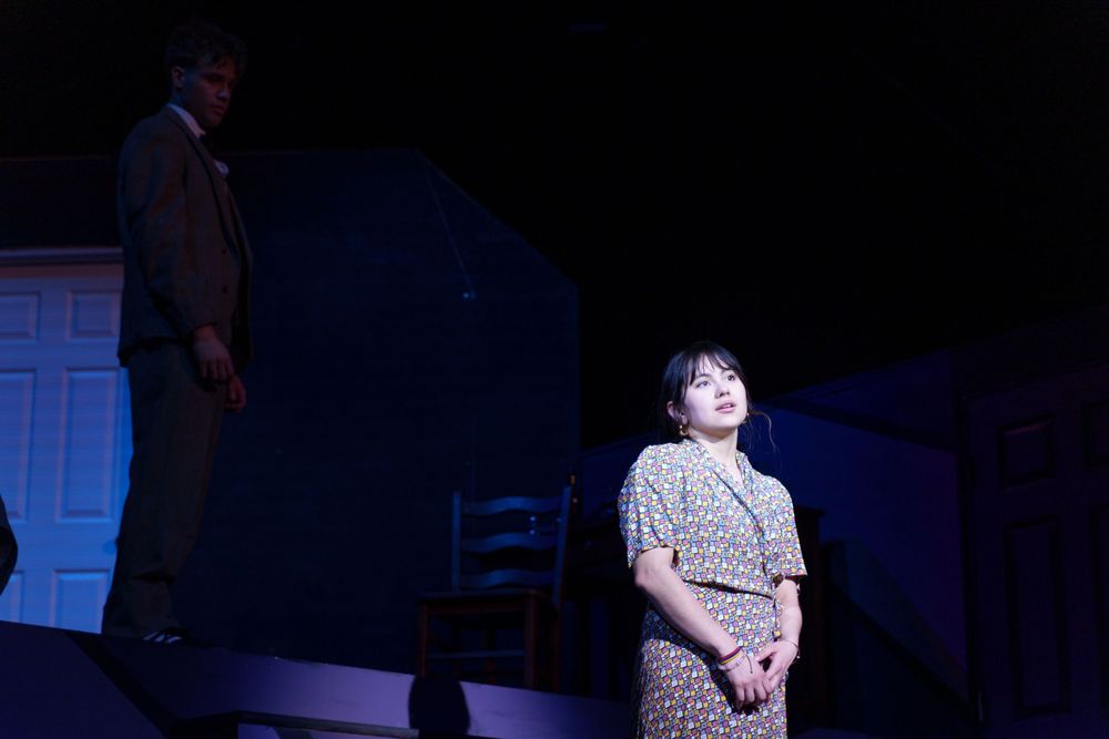 A dramatic stage performance features two actors in a darkly lit theater scene with blue lighting effects.