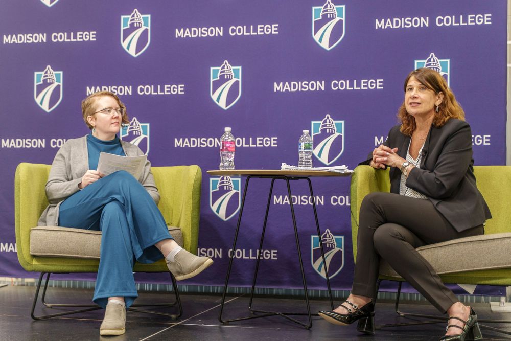 Two people sit in modern chairs having a conversation on stage with Madison College branded backdrop panels.