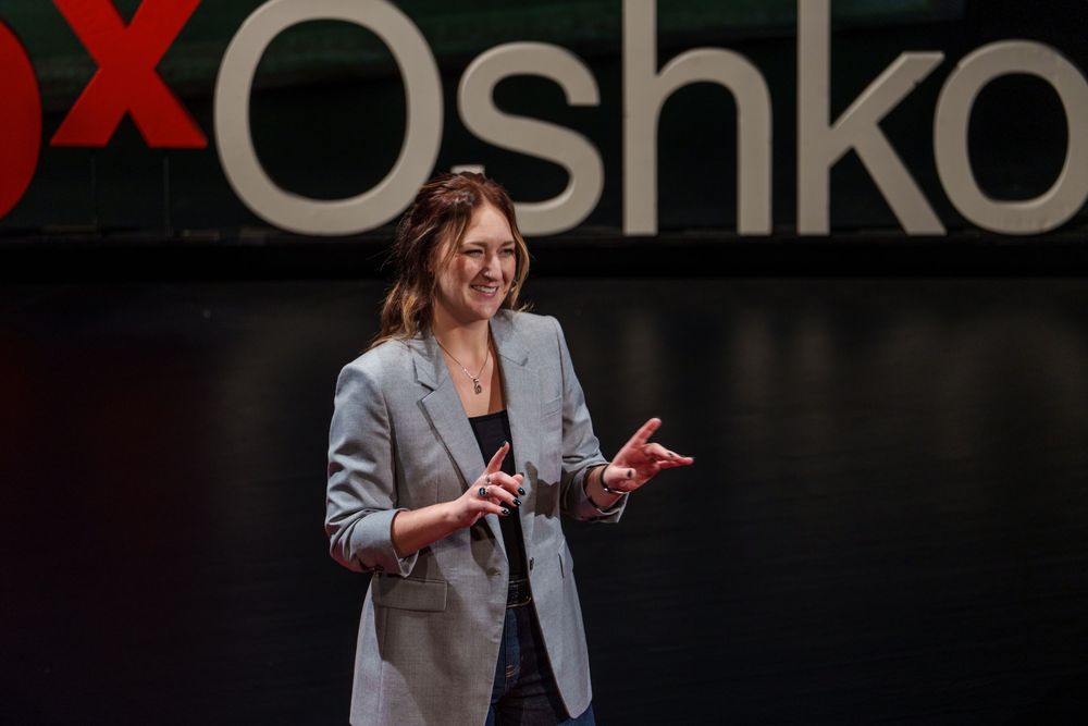 A speaker in a gray blazer gestures while presenting on stage at a TEDx Oshkosh event.