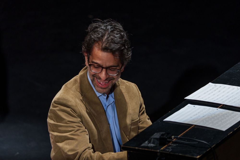 A pianist in a tan jacket and glasses sits at a piano with sheet music on a dark background.