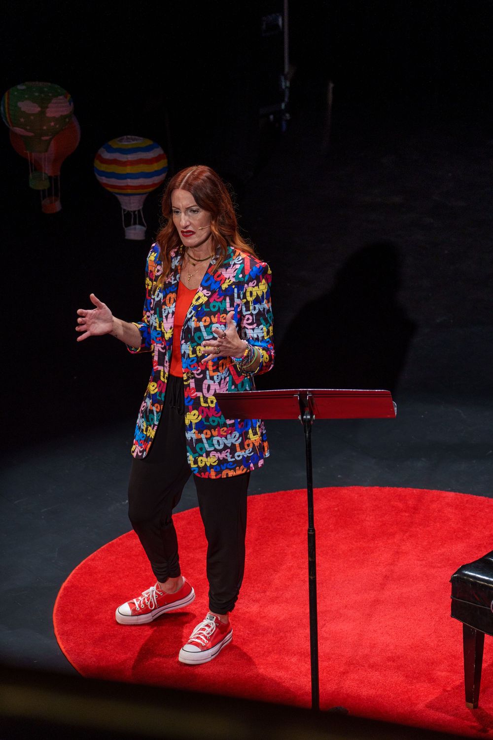 Performer in colorful patterned blazer gestures while standing on a round red carpet near a music stand on dark stage.