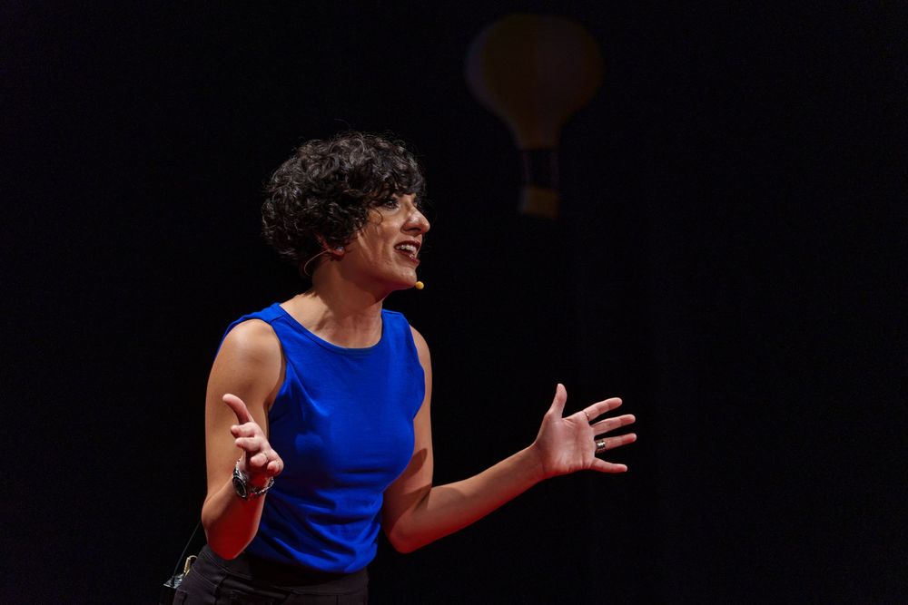 A person in a blue sleeveless top gestures expressively while speaking against a black background.