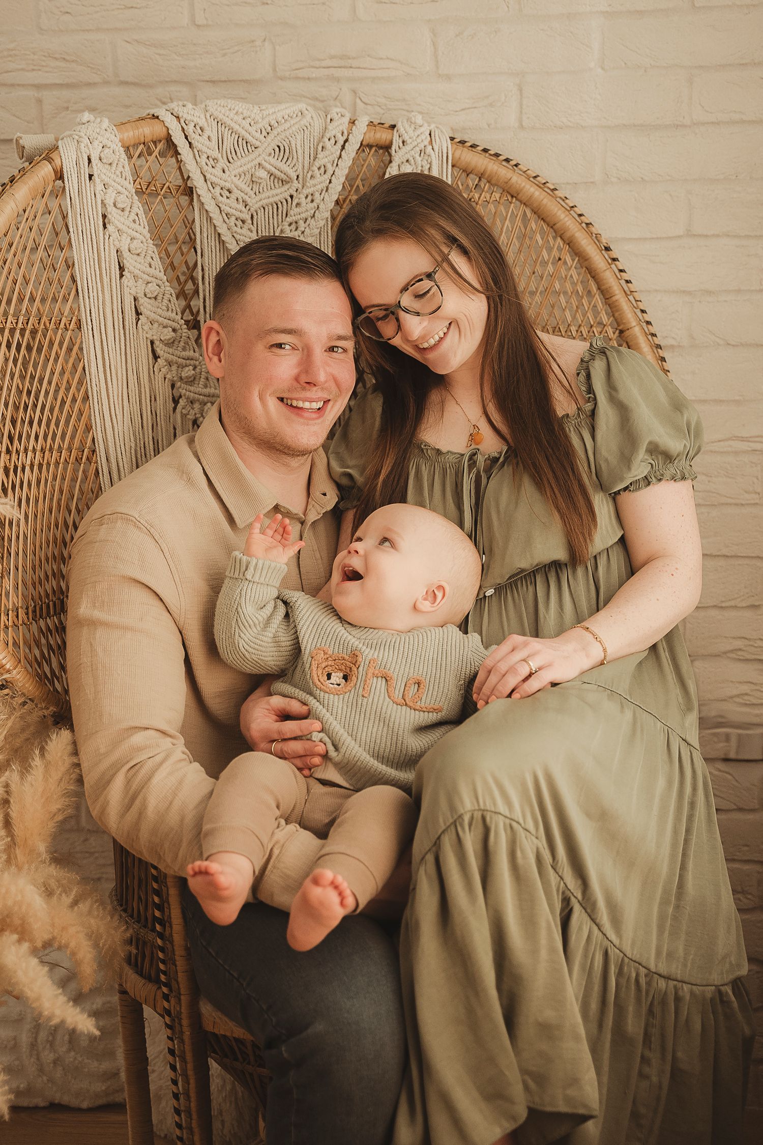 A family sits together in a wicker peacock chair against a brick wall, wearing coordinating neutral-toned outfits.