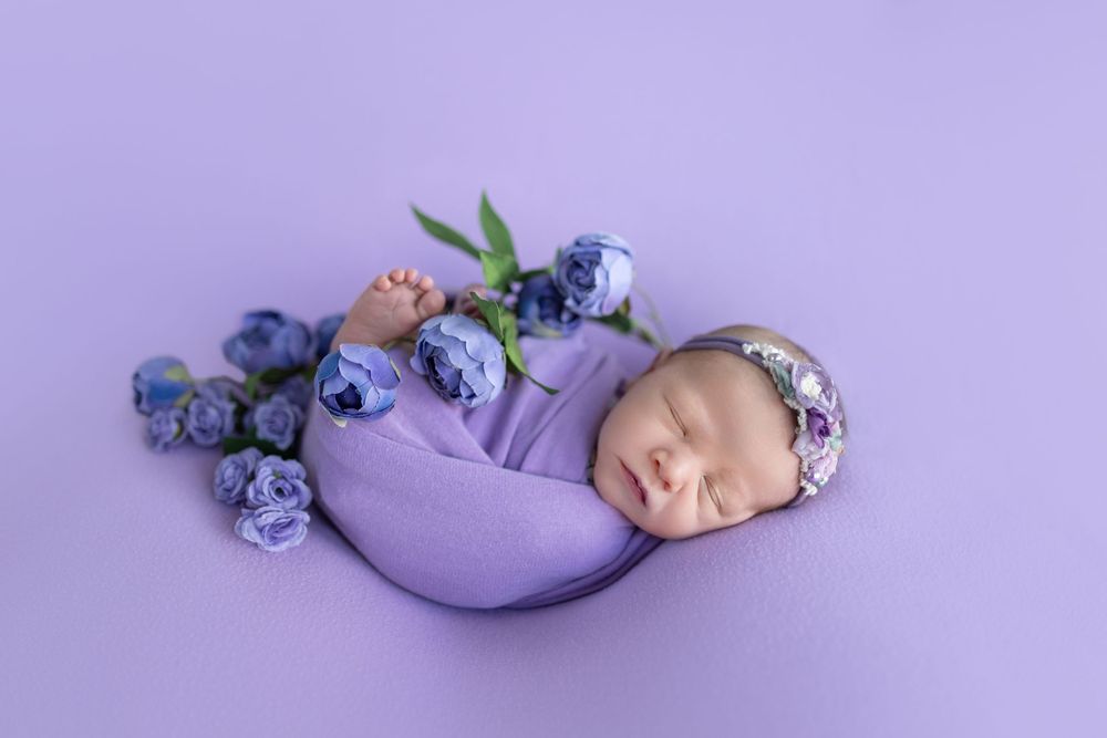 Sleeping newborn on purple backdrop with blue flowers and delicate headband.