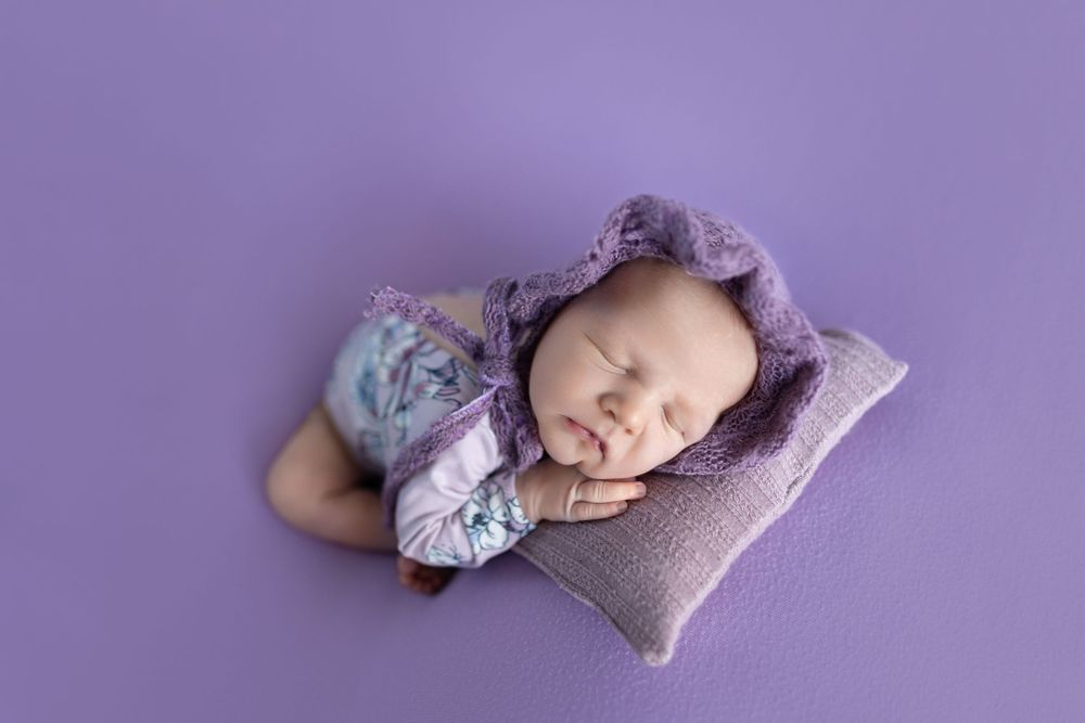 Peaceful newborn sleeping on purple pillow wearing floral outfit.