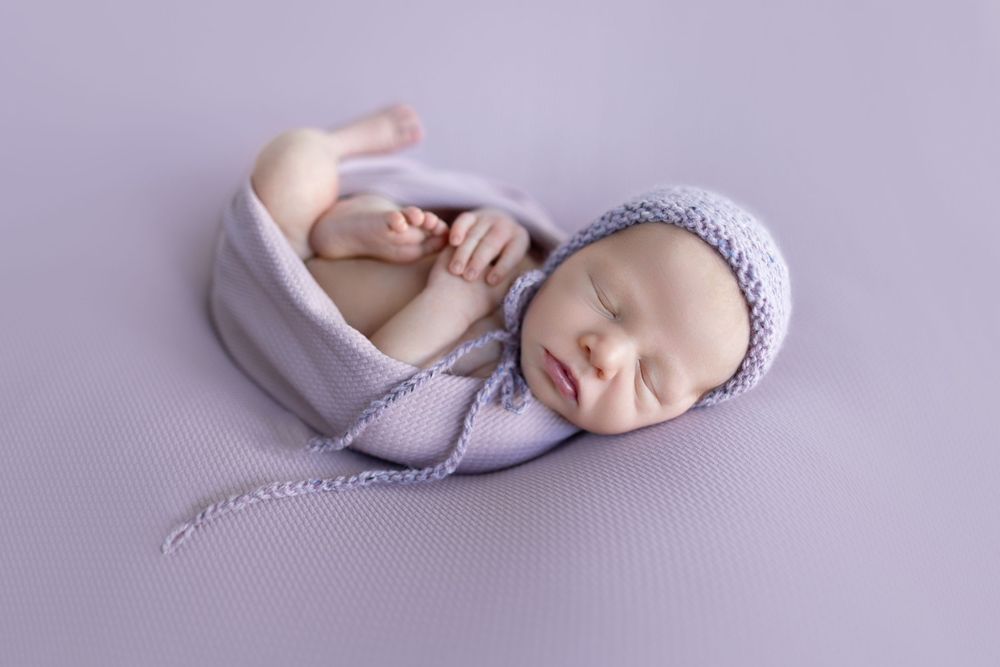 Sleeping newborn wearing knit bonnet on soft purple backdrop in gentle posed portrait.