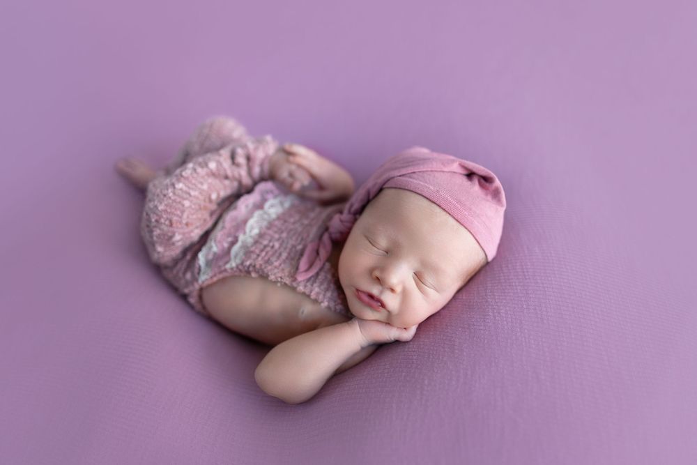 Sleeping baby wearing pink outfit on purple backdrop.