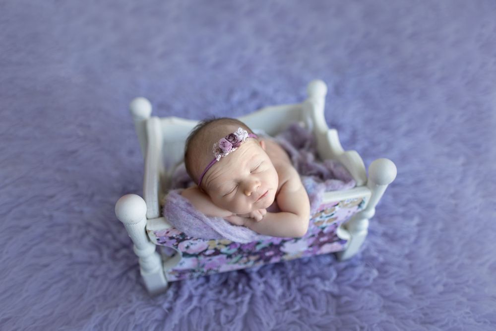 Sleeping newborn on miniature white bed with purple accents and floral headband.