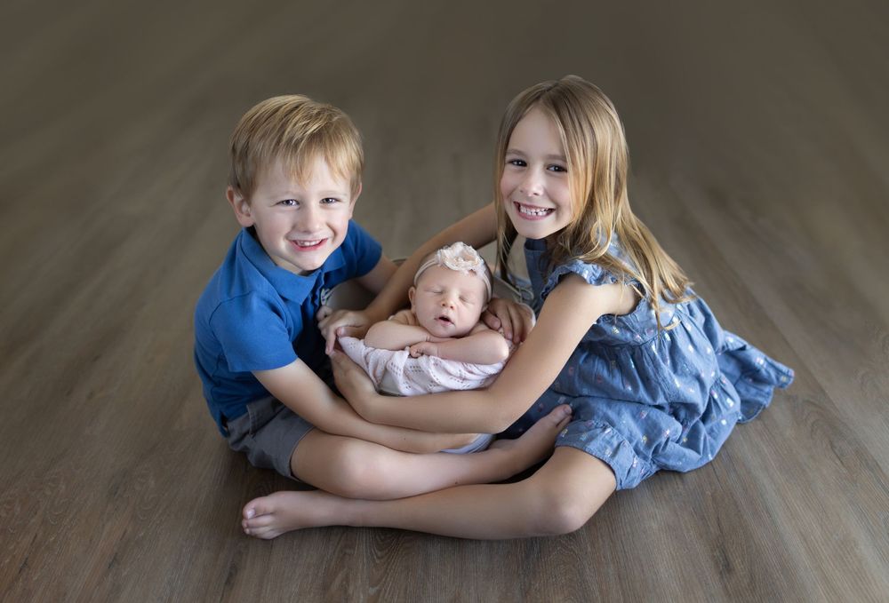 Three siblings in blue and denim outfits sitting together on wooden floor.