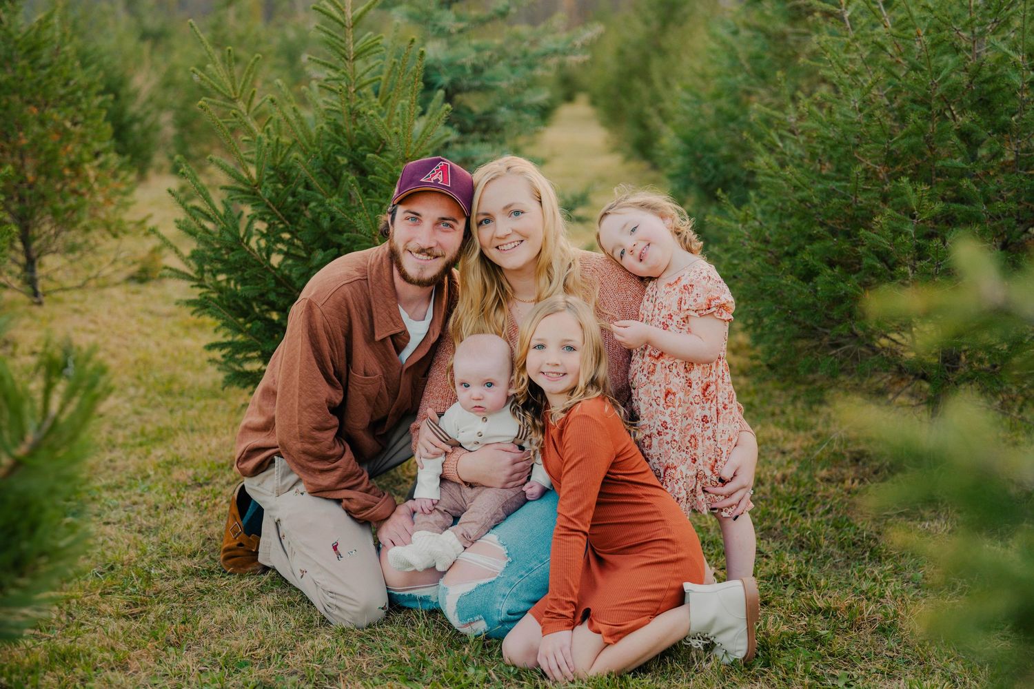 Family poses together at Christmas tree farm surrounded by evergreen trees on a sunny day.