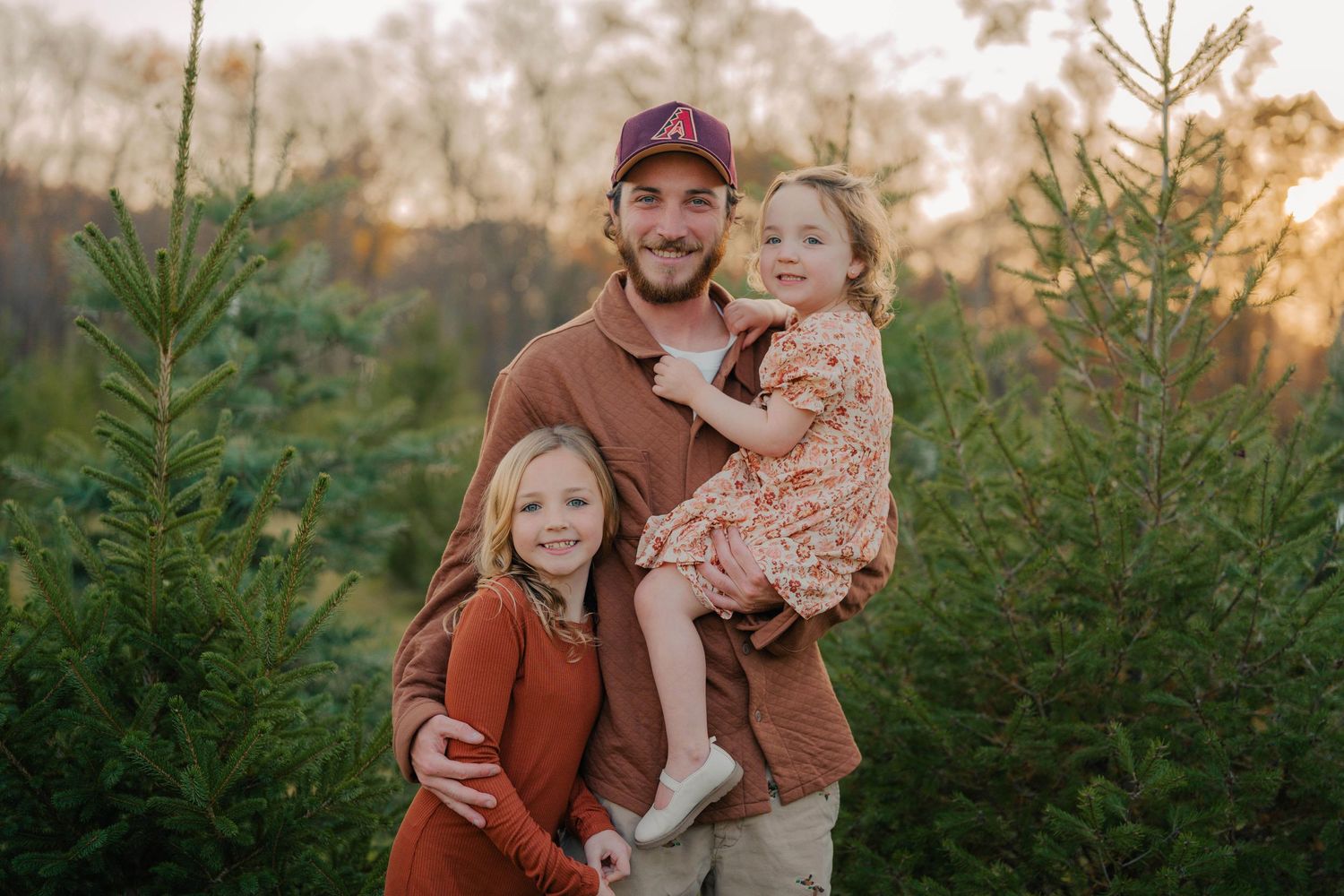 A family in warm autumn clothing poses together at a Christmas tree farm during golden hour sunset.
