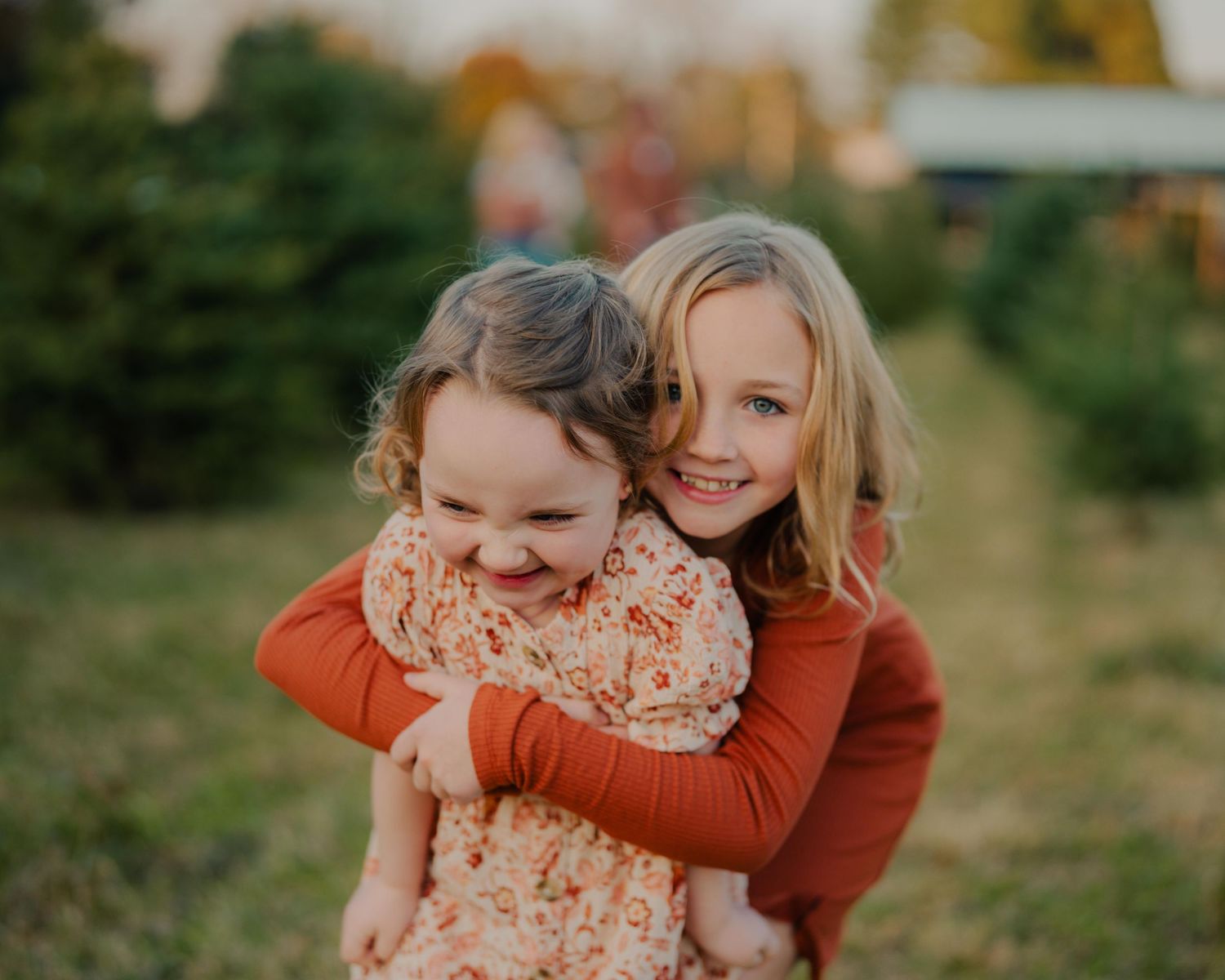 Two children in orange and coral outfits smile and embrace while playing outdoors during autumn.