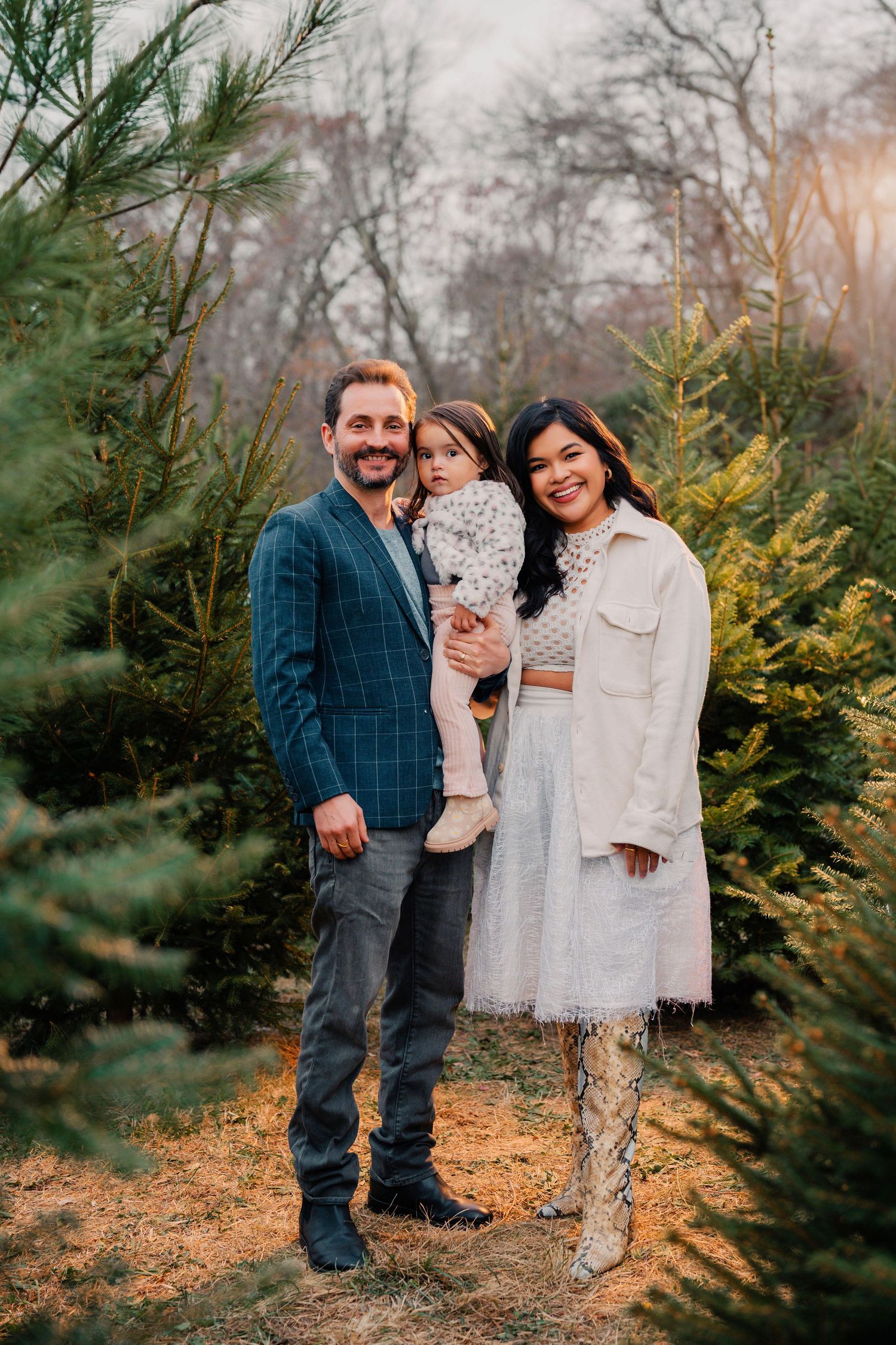 A family poses together outdoors among evergreen trees during an autumn photo session at a Christmas tree farm.