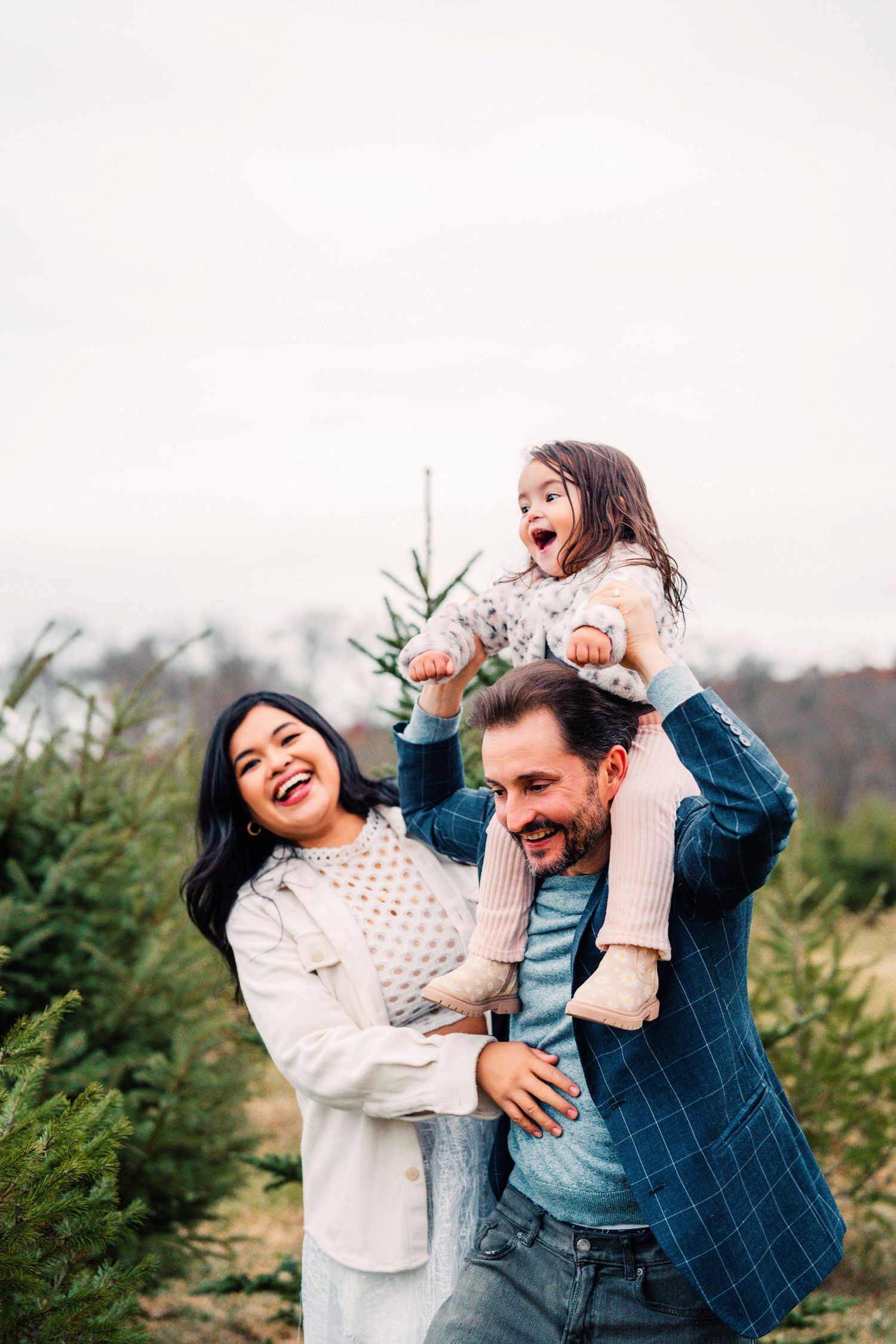 Joyful family laughs together while spending time at a Christmas tree farm on an overcast winter day.