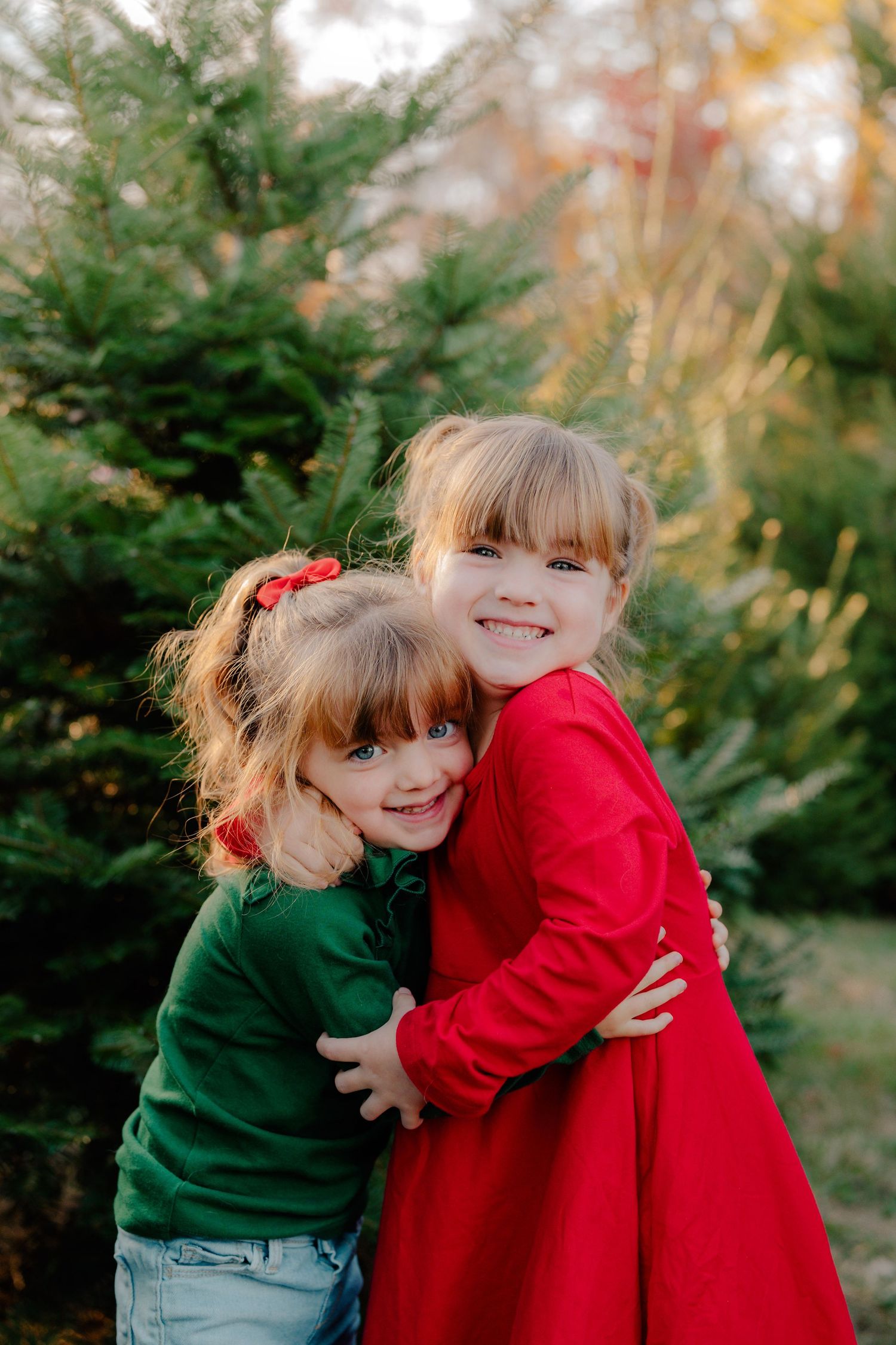 Two children wearing festive red and green sweaters embrace while smiling in front of evergreen trees.